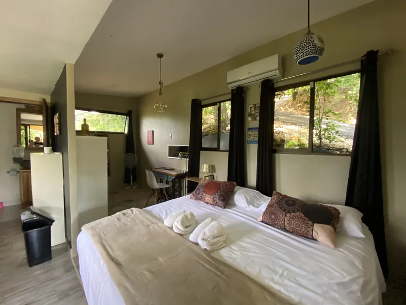 The studio bedroom with a king bed, folded towels, a work desk, and garden windows dressed in black curtains.