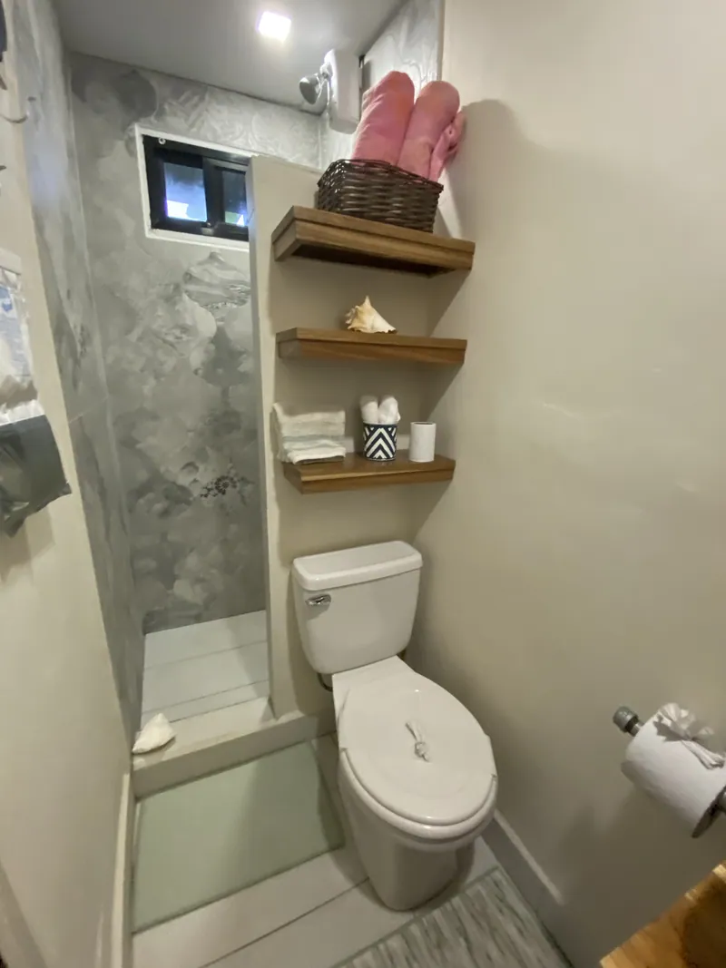 The compact bathroom with a walk-in tiled shower, floating wood shelves, and rolled towels in a basket.